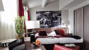 A modern hotel room with a wooden desk, a laptop, a red chair, a bed with red and white bedding, a large window with curtains, and a wall-mounted TV. - Aparthotel Adagio Aix en Provence Centre (Aix-en-Provence)