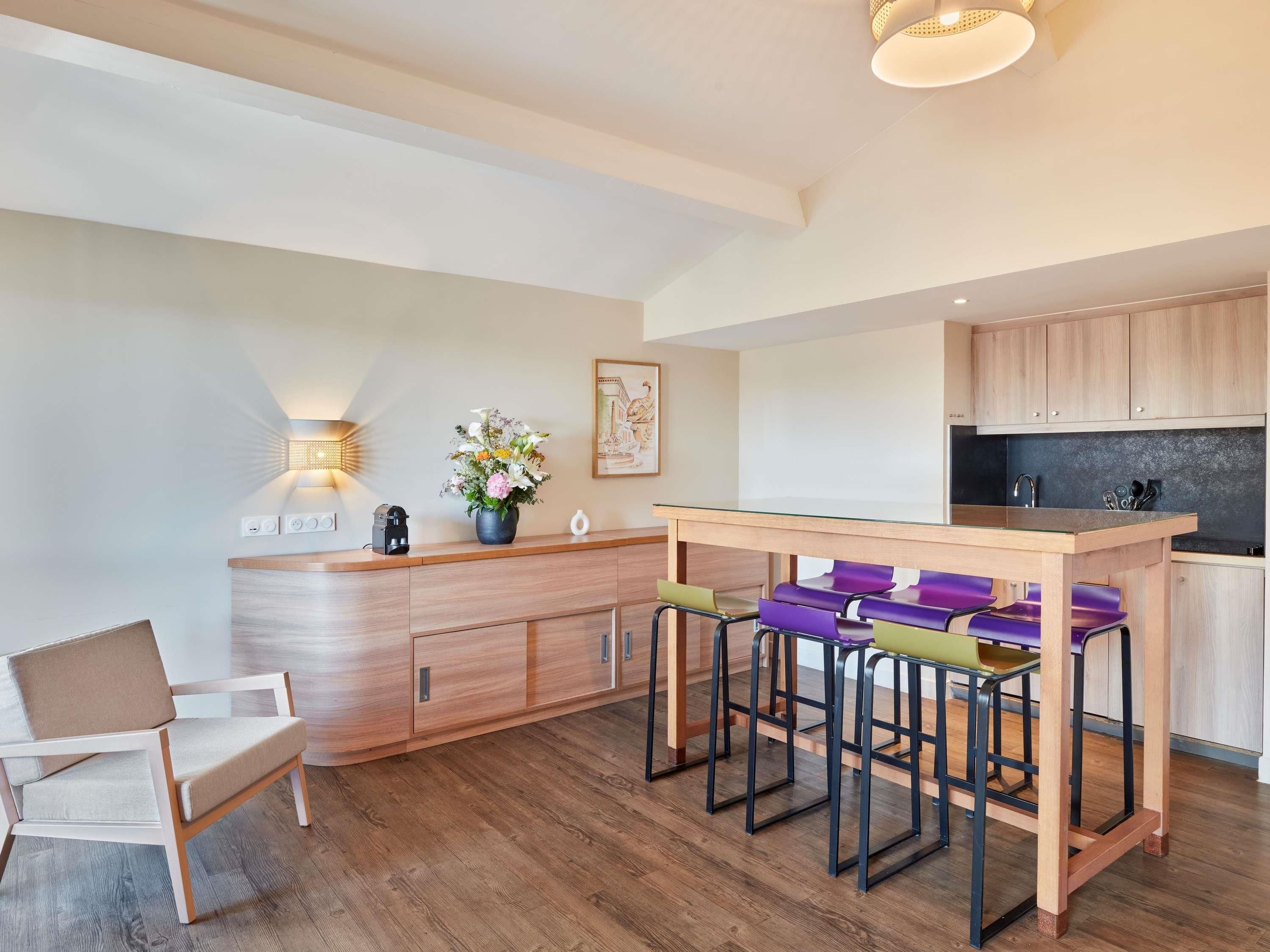 Una cocina moderna con armarios de madera, una isla central con taburetes altos, una lámpara de pared y una pequeña mesa con un jarrón de flores.