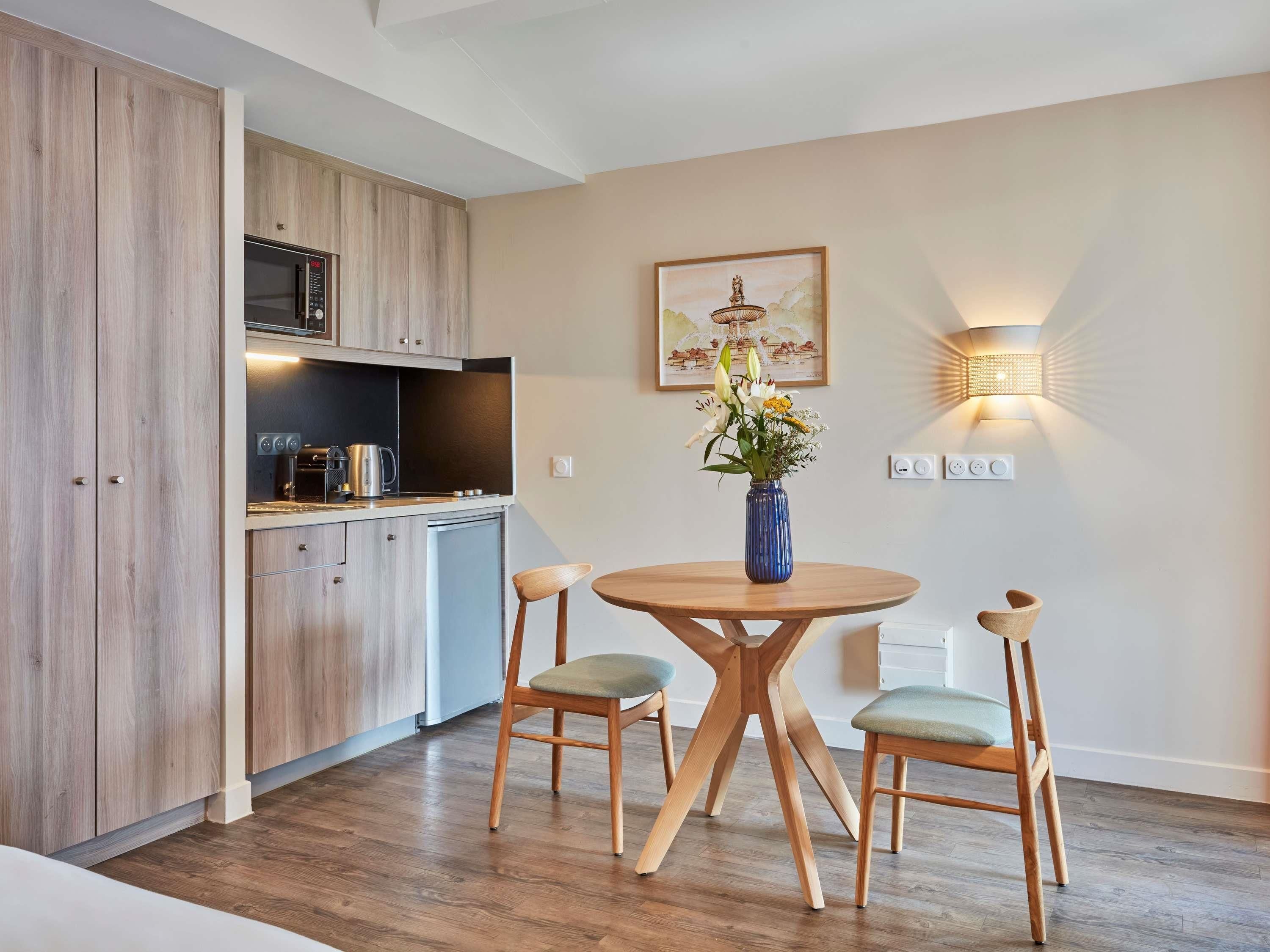 Una cocina moderna con armarios de madera, una mesa de comedor con dos sillas, un jarrón con flores y una lámpara de pared.