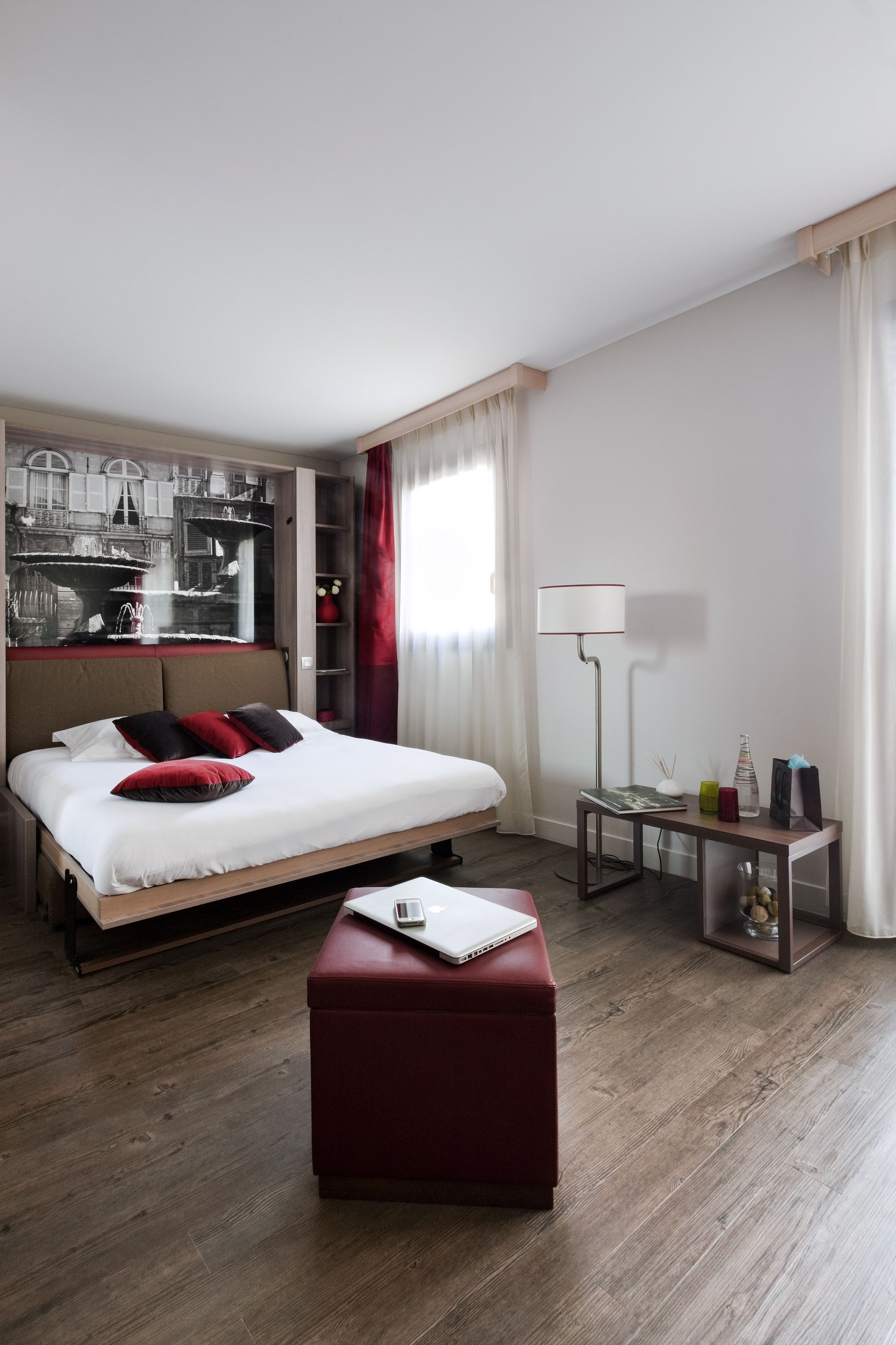 Een moderne hotelkamer met een groot bed, een rode poef, een bureau met een lamp en een stadsgezicht kunstwerk aan de muur.