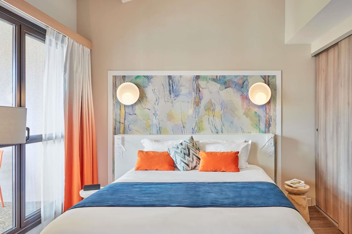 A modern bedroom with a large bed, colorful artwork on the headboard, and a view of the outdoors through a large window.