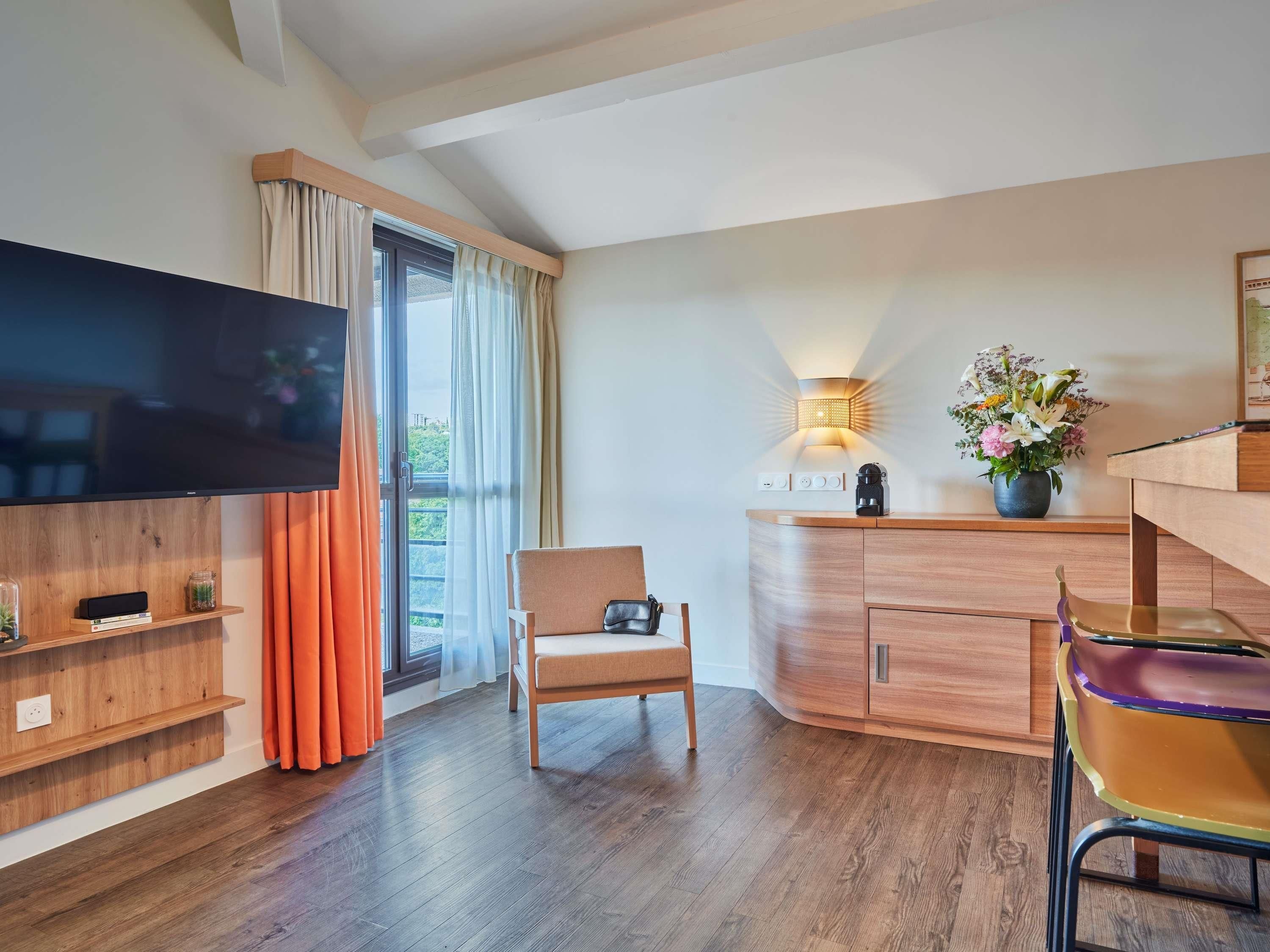 Une chambre moderne dotée d’une télévision à écran plat, de meubles en bois, d’un coin repas et d’une fenêtre avec des rideaux.