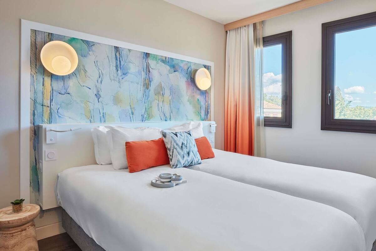 A hotel room with a large bed, a headboard with abstract art, two bedside lamps, and a view of the outdoors through a window.
