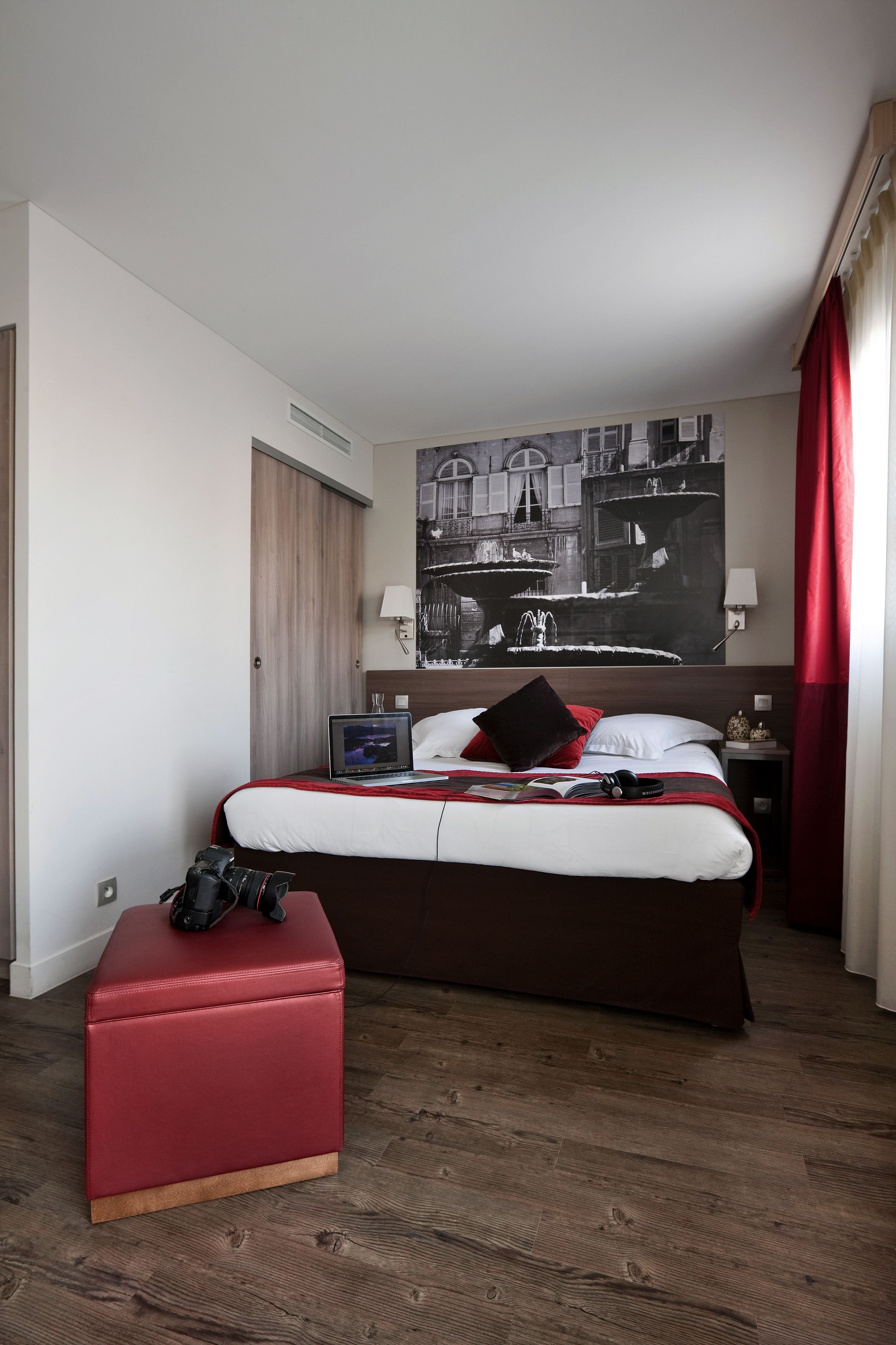 A hotel room with a large bed, a red ottoman, a laptop on the bed, and a black and white artwork on the wall.