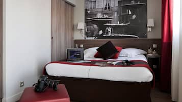 Ein Hotelzimmer mit einem großen Bett, einem roten Hocker, einem Laptop auf dem Bett und einem Schwarz-Weiß-Kunstwerk an der Wand.