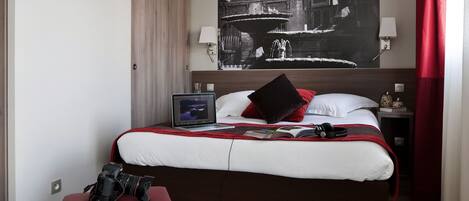 A hotel room with a large bed, a red ottoman, a laptop on the bed, and a black and white artwork on the wall.