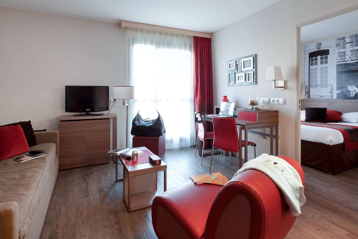 A modern hotel room with a sofa, a desk with a laptop, a television, and a bed with red bedding.