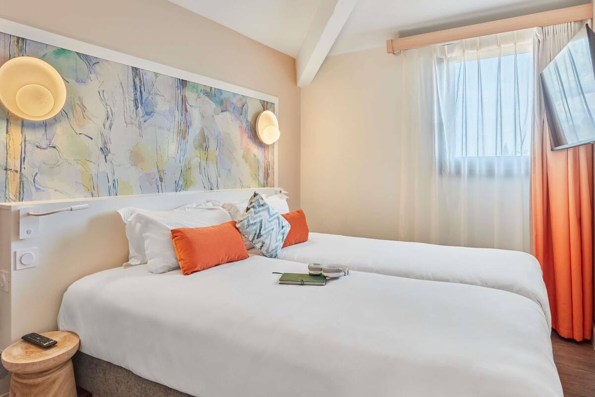 A hotel room with a large bed, a bedside table, a wall-mounted light, and a window with curtains.