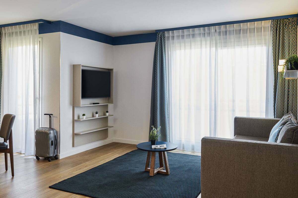 A modern hotel room with a flat-screen TV, a small table with a plant, a sofa, a suitcase, and a window with sheer curtains.
