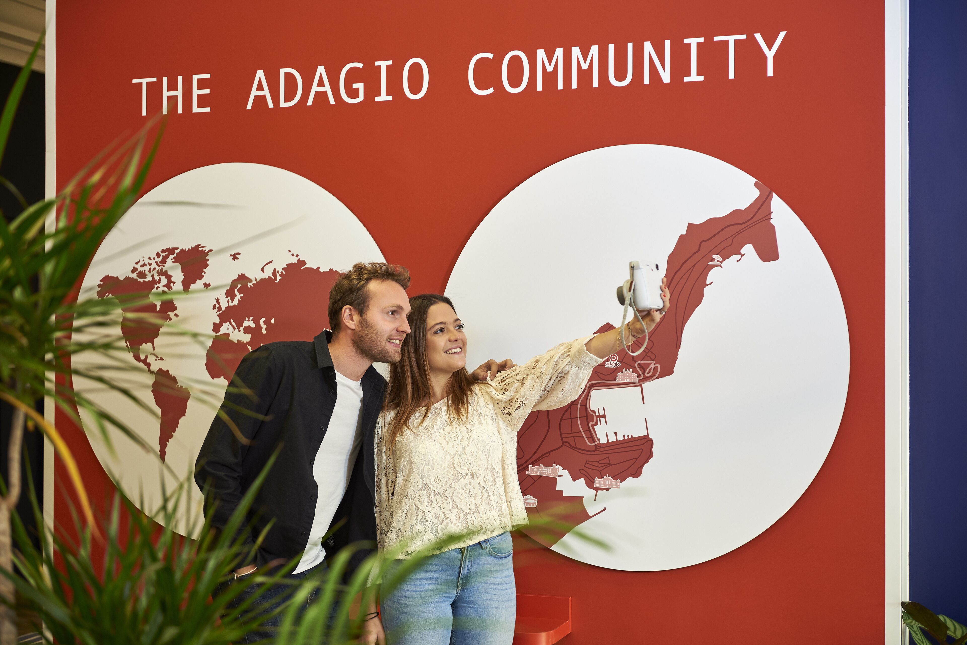 Una pareja tomándose un selfie frente a un muro con un mapa y el texto “THE ADAGIO COMMUNITY”.