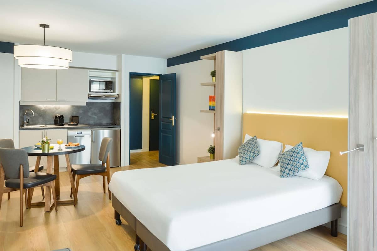 A modern hotel room with a bed, bedside table, and a kitchenette.