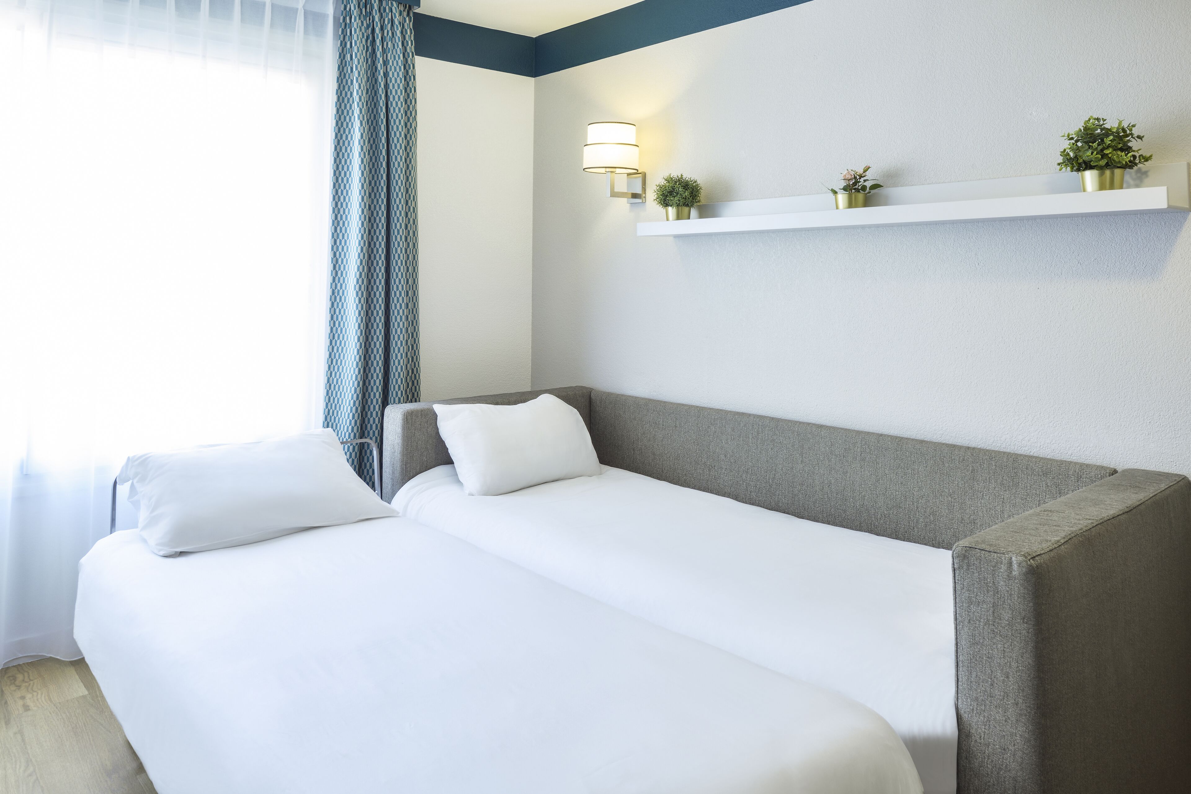 Habitación de hotel con cama, mesita de noche, estante en la pared con plantas y ventana con cortinas.