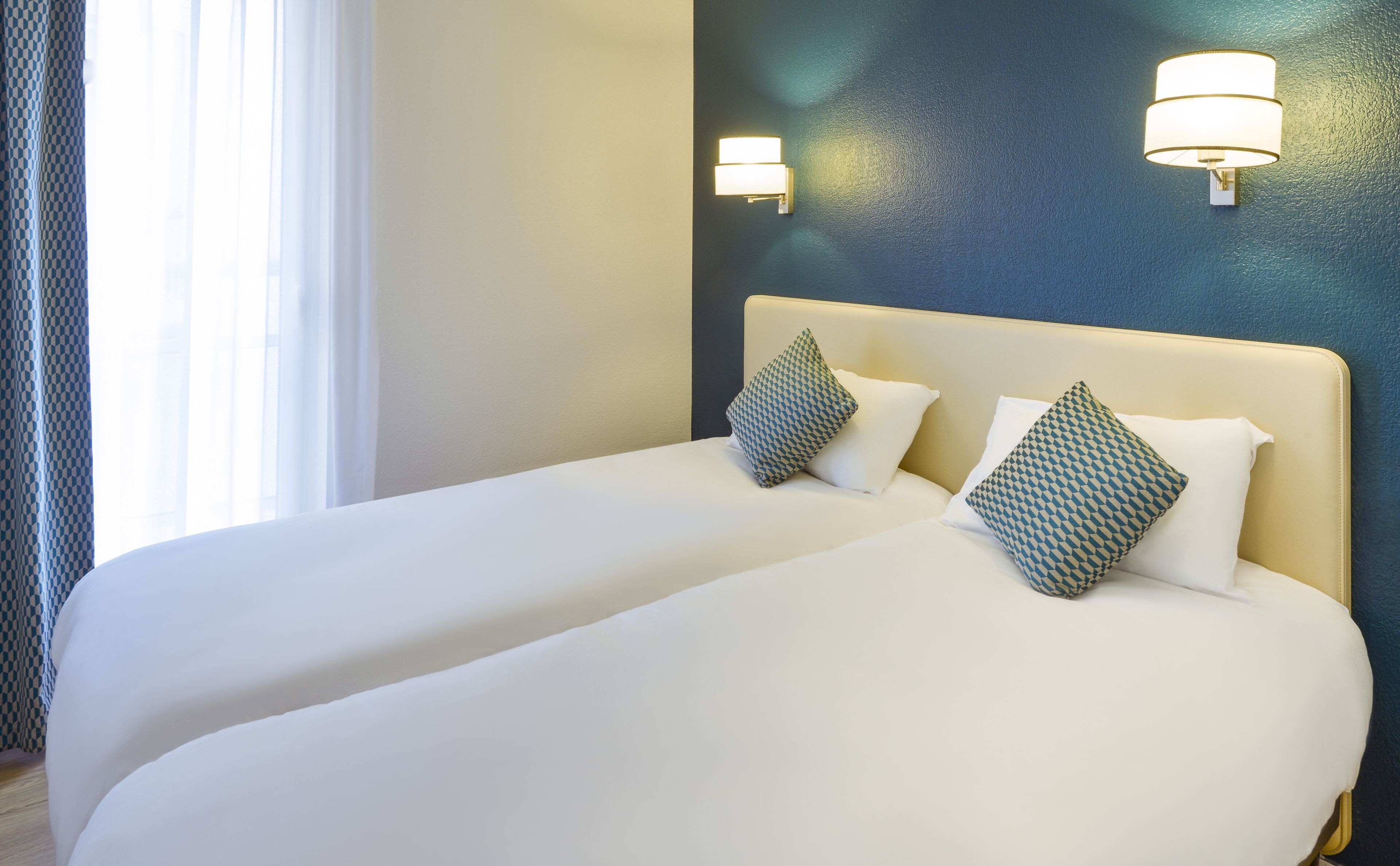 Une chambre d’hôtel avec deux lits, chacun agrémenté de draps blancs et d’oreillers à motifs, un mur d’accent bleu et des luminaires fixés au mur.