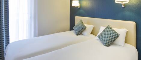 A hotel room with two beds, each with white bedding and patterned pillows, a blue accent wall, and wall-mounted lighting.