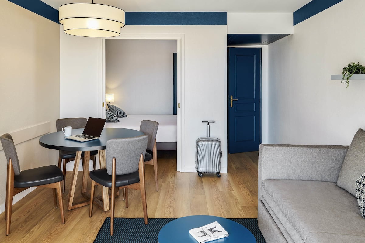 A modern hotel room with a dining area, a sofa, a small table with a magazine, a lamp, a suitcase, and a door.