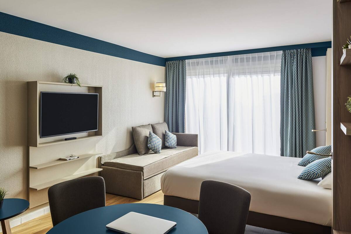 A modern hotel room with a flat-screen TV, a sofa, a bed with patterned pillows, a desk with a laptop, and a large window with sheer curtains.