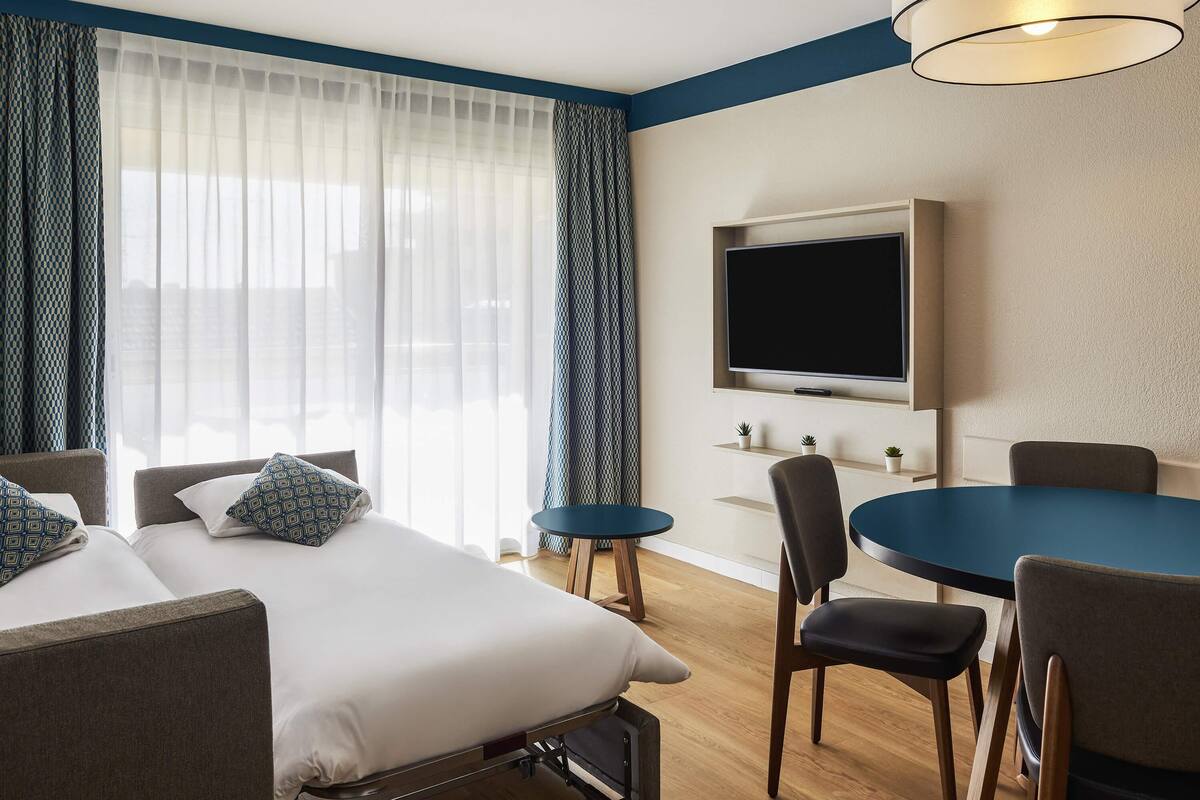 A hotel room with a bed, a desk, a chair, a TV, and a window with curtains.