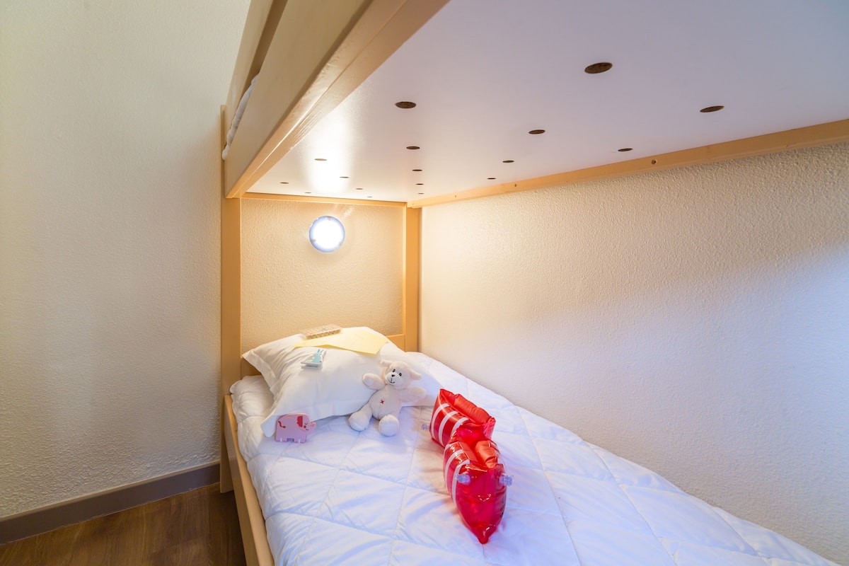 Een kleine kamer met een enkel bed, een houten hoofdbord, een wit matras, een wit kussen, een pluchen speeltje en een rode decoratie.