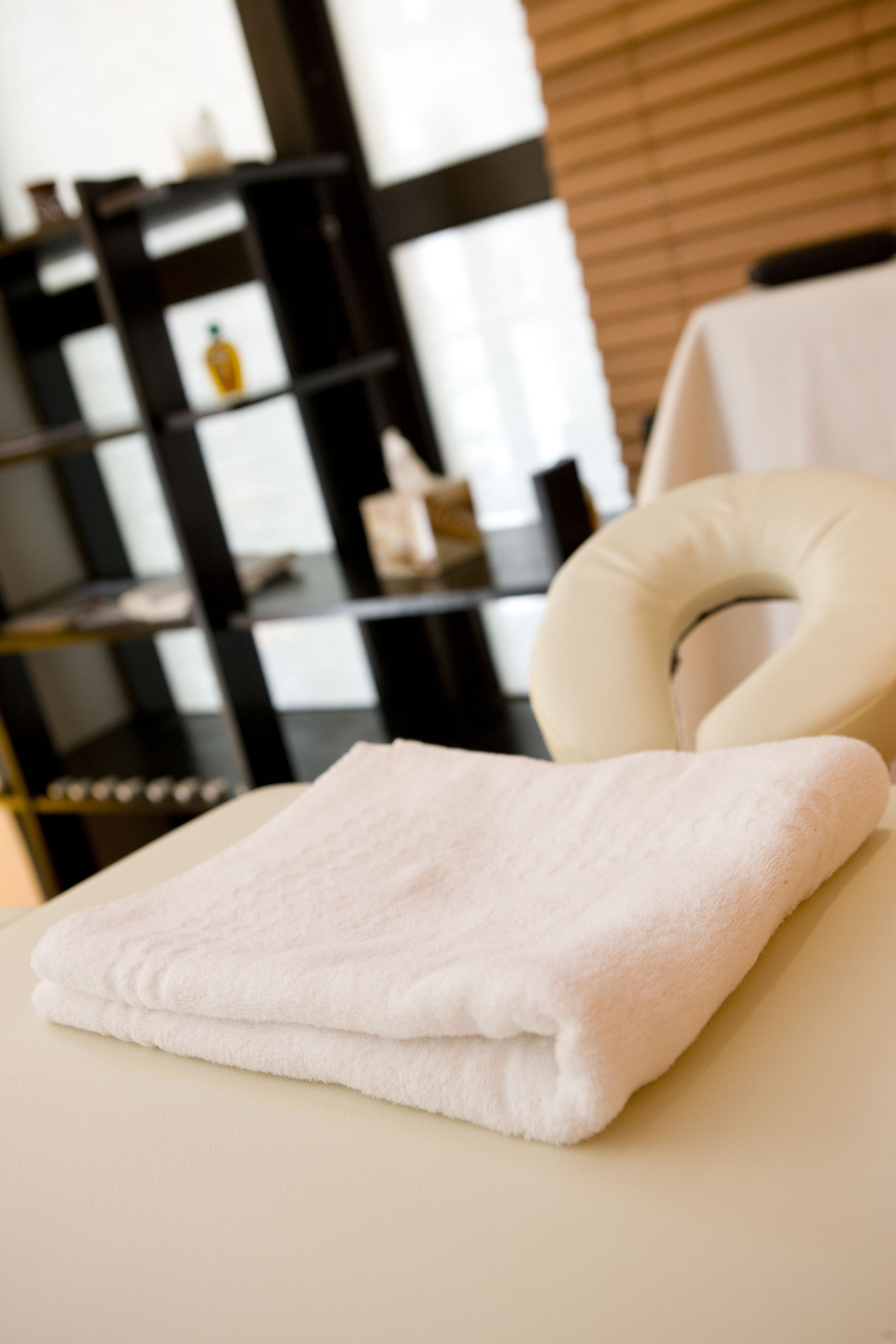 Une serviette blanche, soigneusement pliée, est posée sur une table de massage, avec un coussin pour la nuque. La pièce est dotée d’un mur en bois et, au fond, on aperçoit une étagère noire.