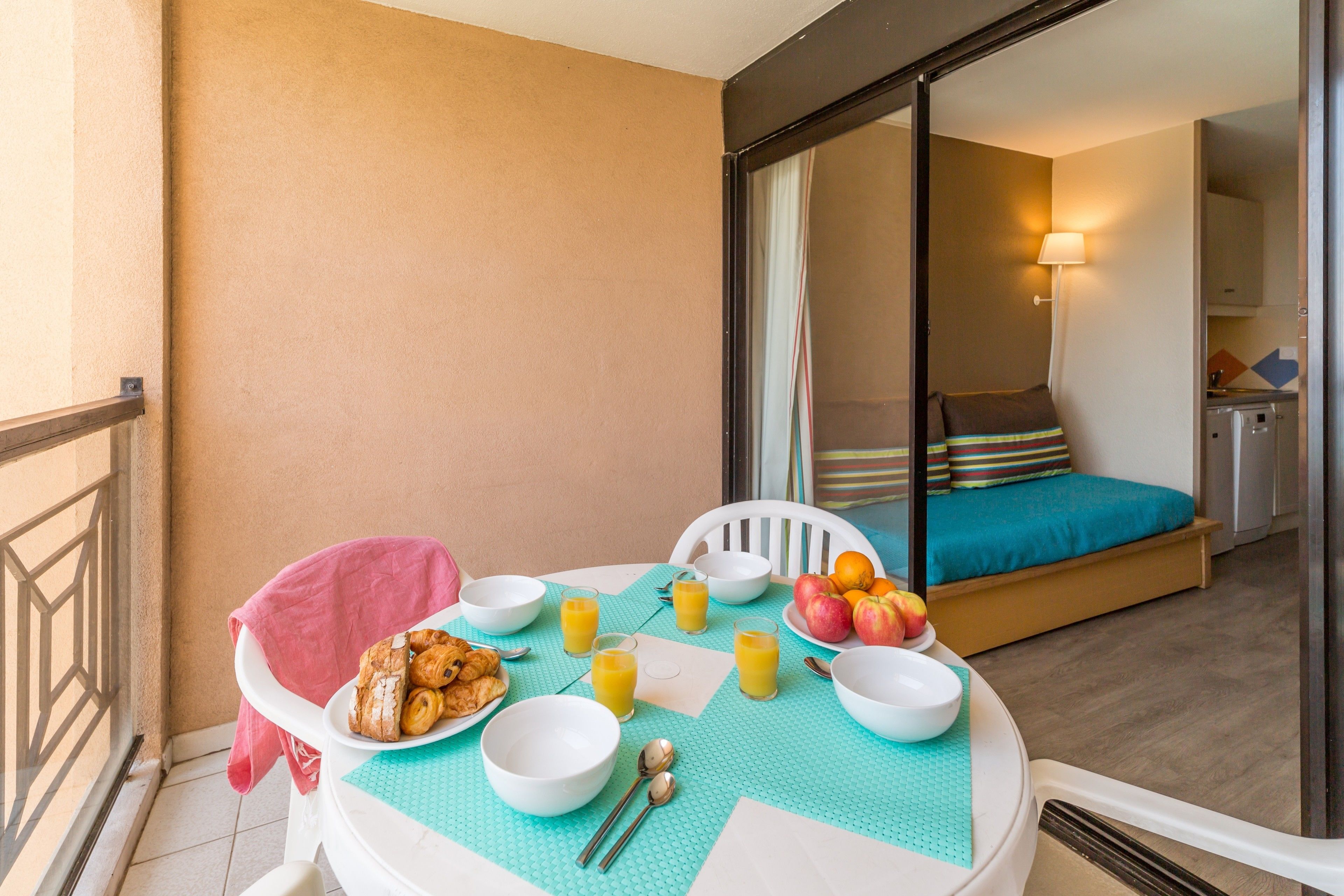 A balcony with breakfast setup including croissants, fruit, and juice, and a bed with a striped pillow and teal bedspread.