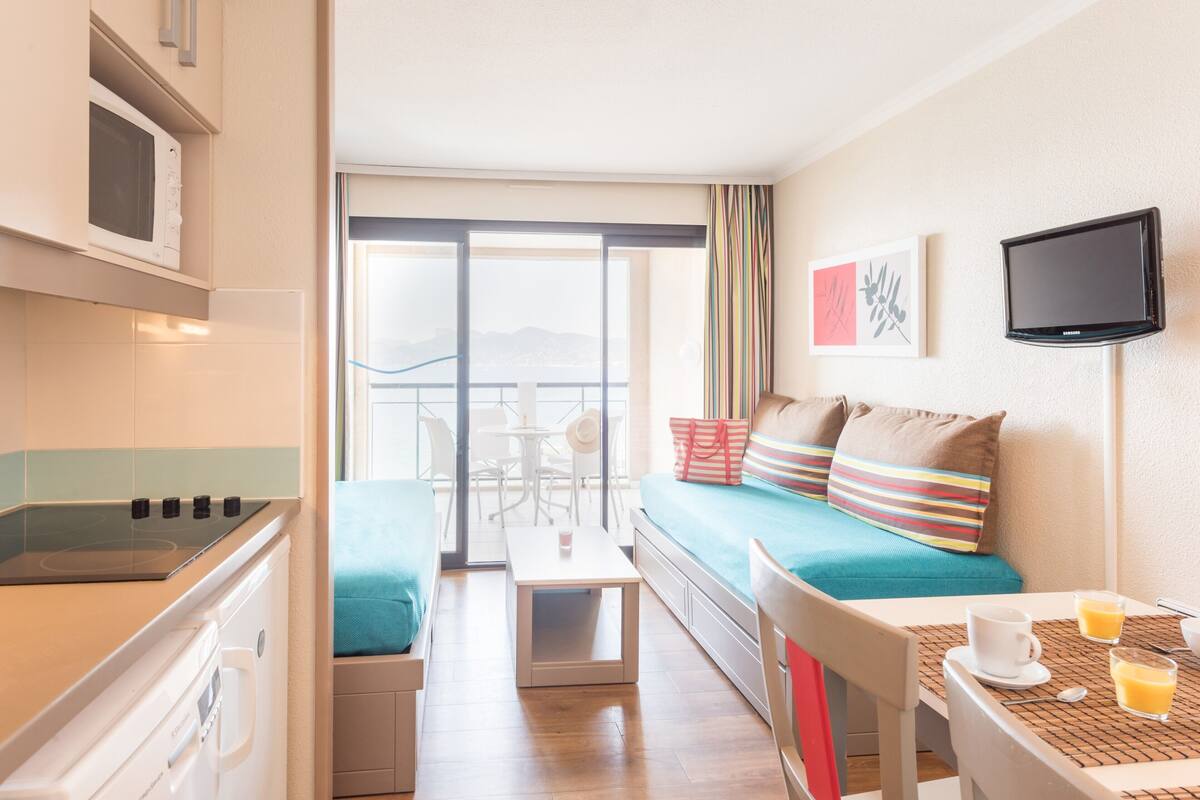 Een compacte hotelkamer met een kitchenette, een balkon met uitzicht, een bank met gestreepte kussens en een televisie aan de muur.