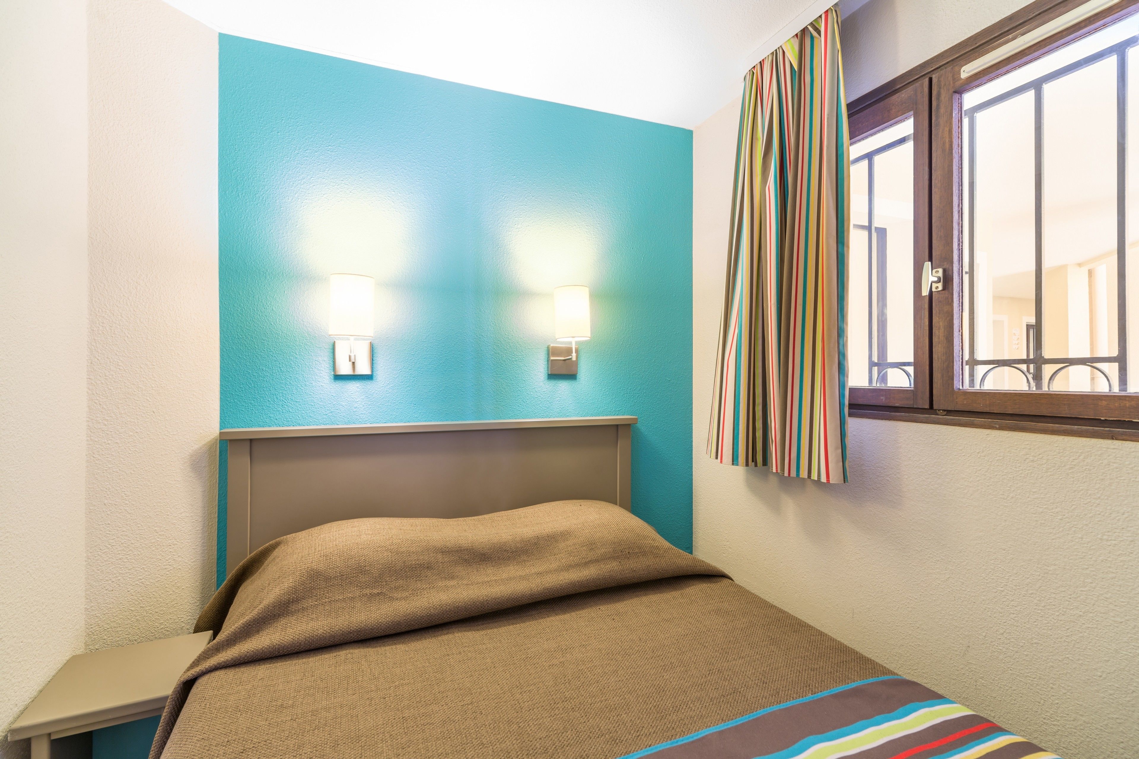 Une chambre à coucher avec un lit, deux lampes fixées au mur, une fenêtre avec des rideaux et une vue sur l’extérieur.