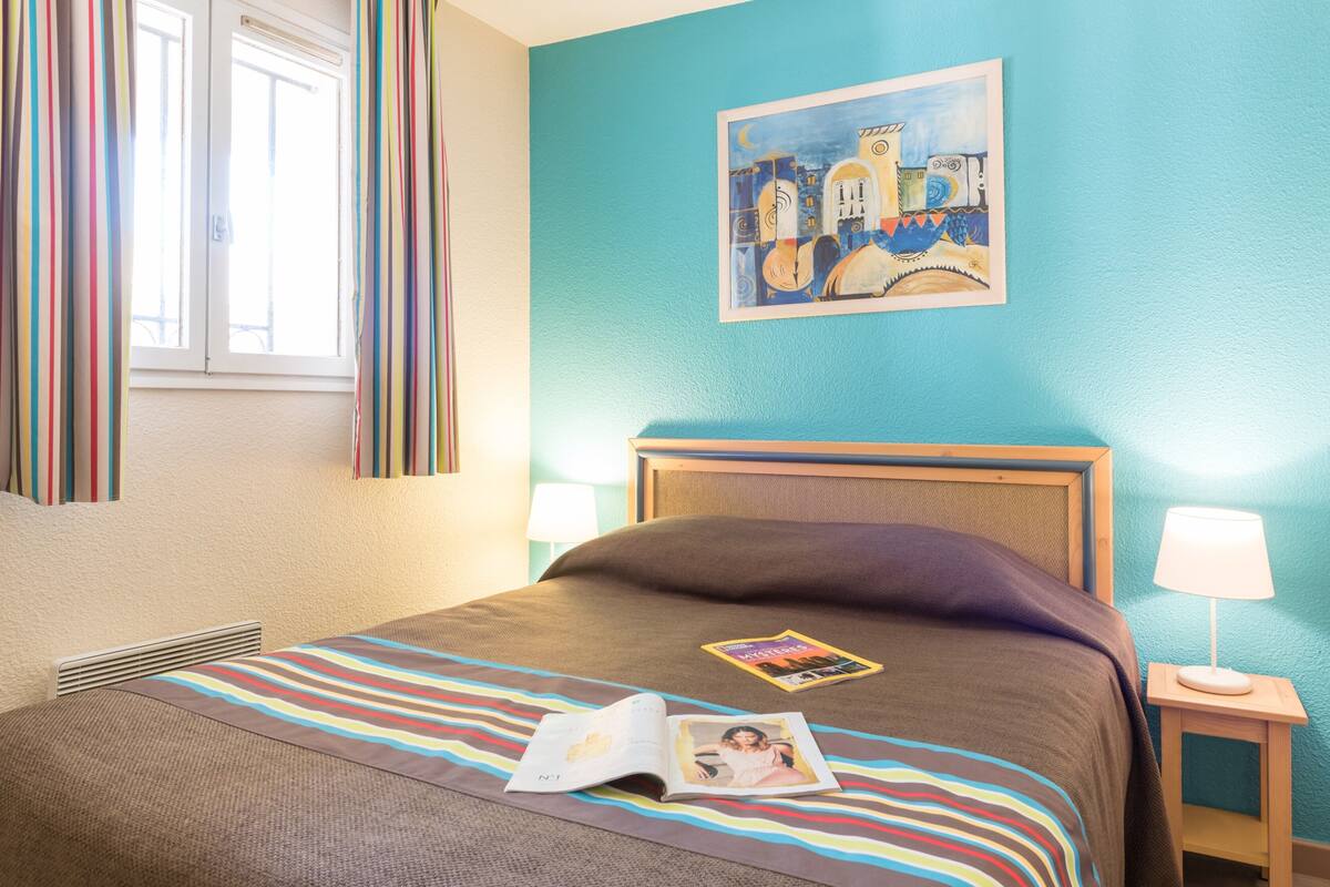Een hotelkamer met een bed, een raam met gordijnen, een schilderij aan de muur, een nachtkastje met een lamp en een tijdschrift op het bed.