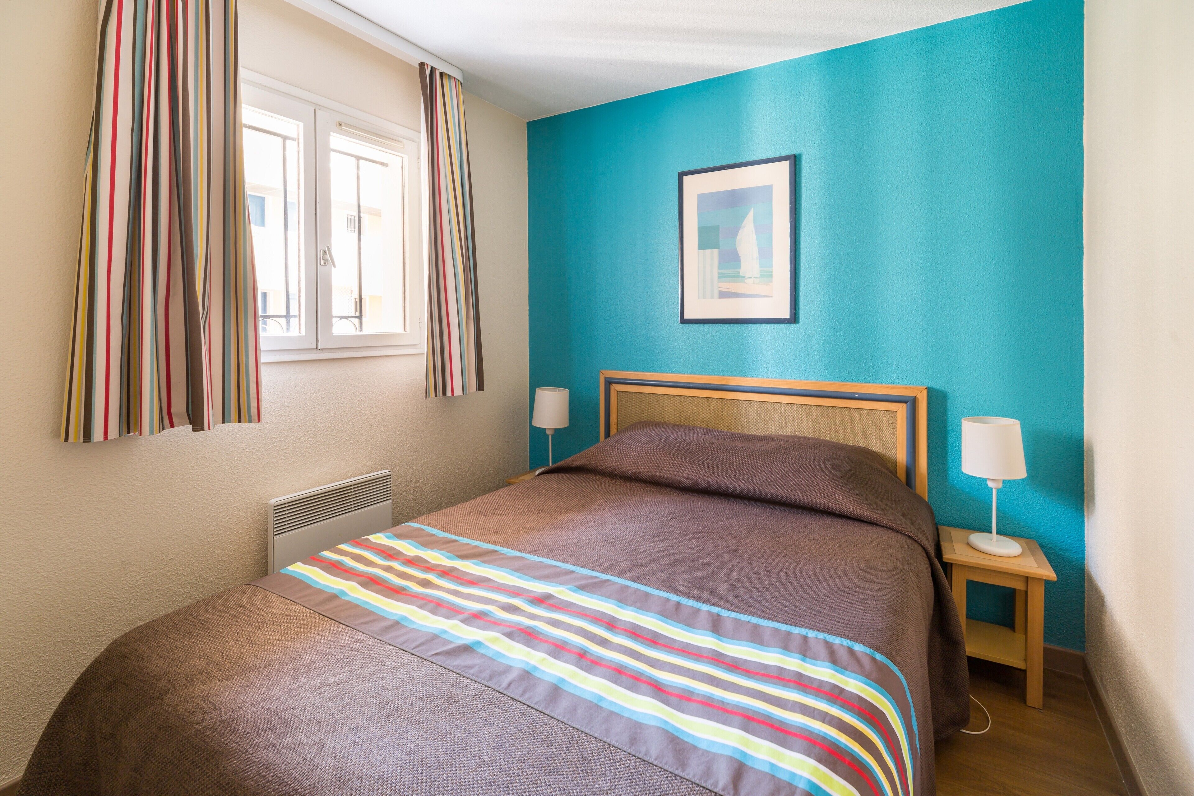 Ein Schlafzimmer mit Bett, einem Fenster mit Vorhängen, einem Nachttisch mit Lampe und einem gerahmten Bild an der Wand.
