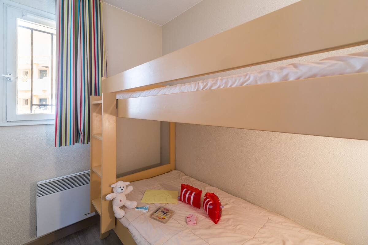 Een slaapkamer met stapelbed, raam, verwarming, een teddybeer en persoonlijke spullen op het bed.