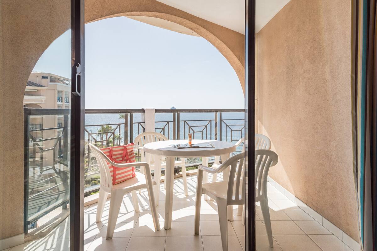 Een balkon met een ronde tafel en stoelen, met uitzicht op water en gebouwen.