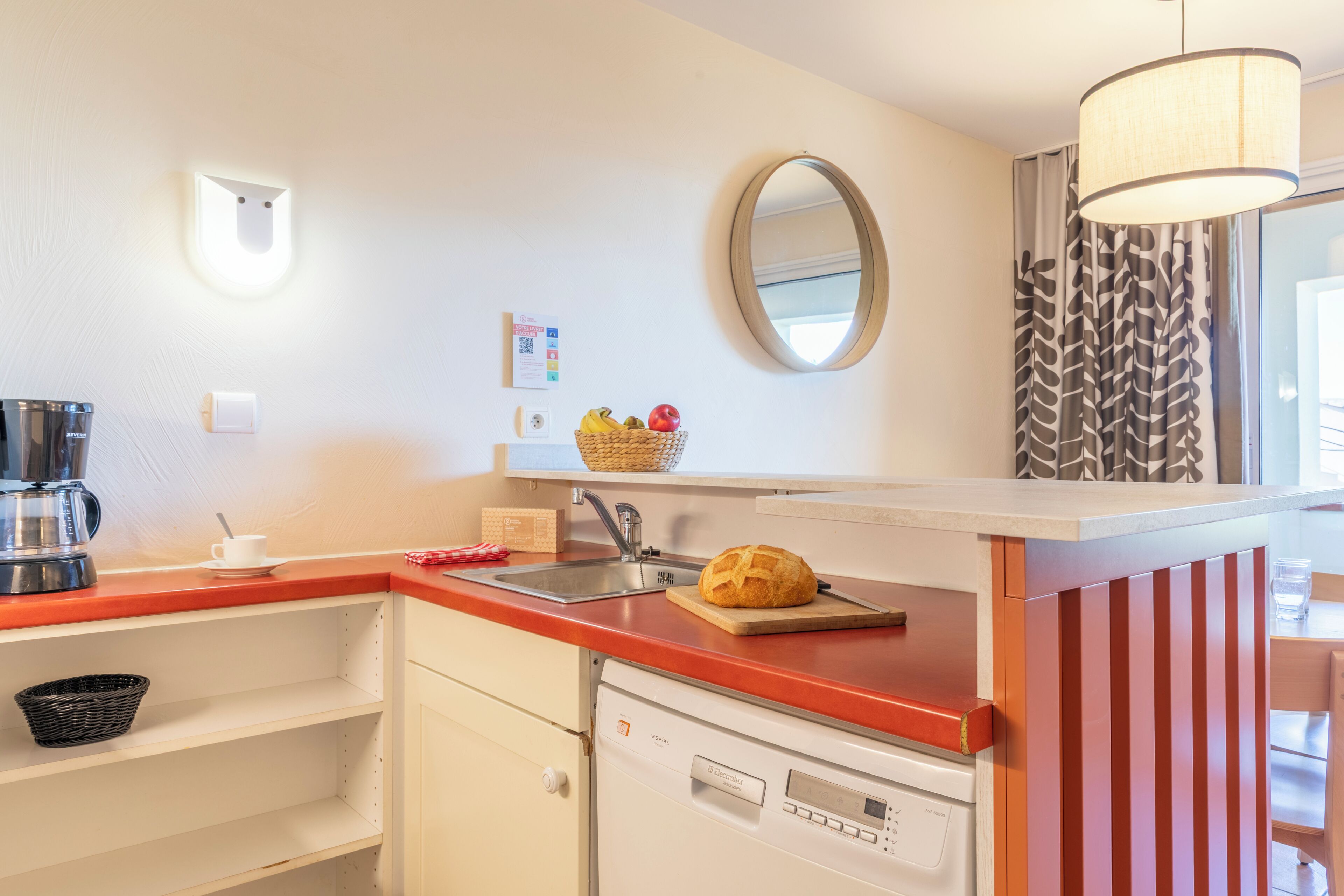 Una cocina con cafetera, hogaza de pan, cesta de fruta, pileta y espejo.