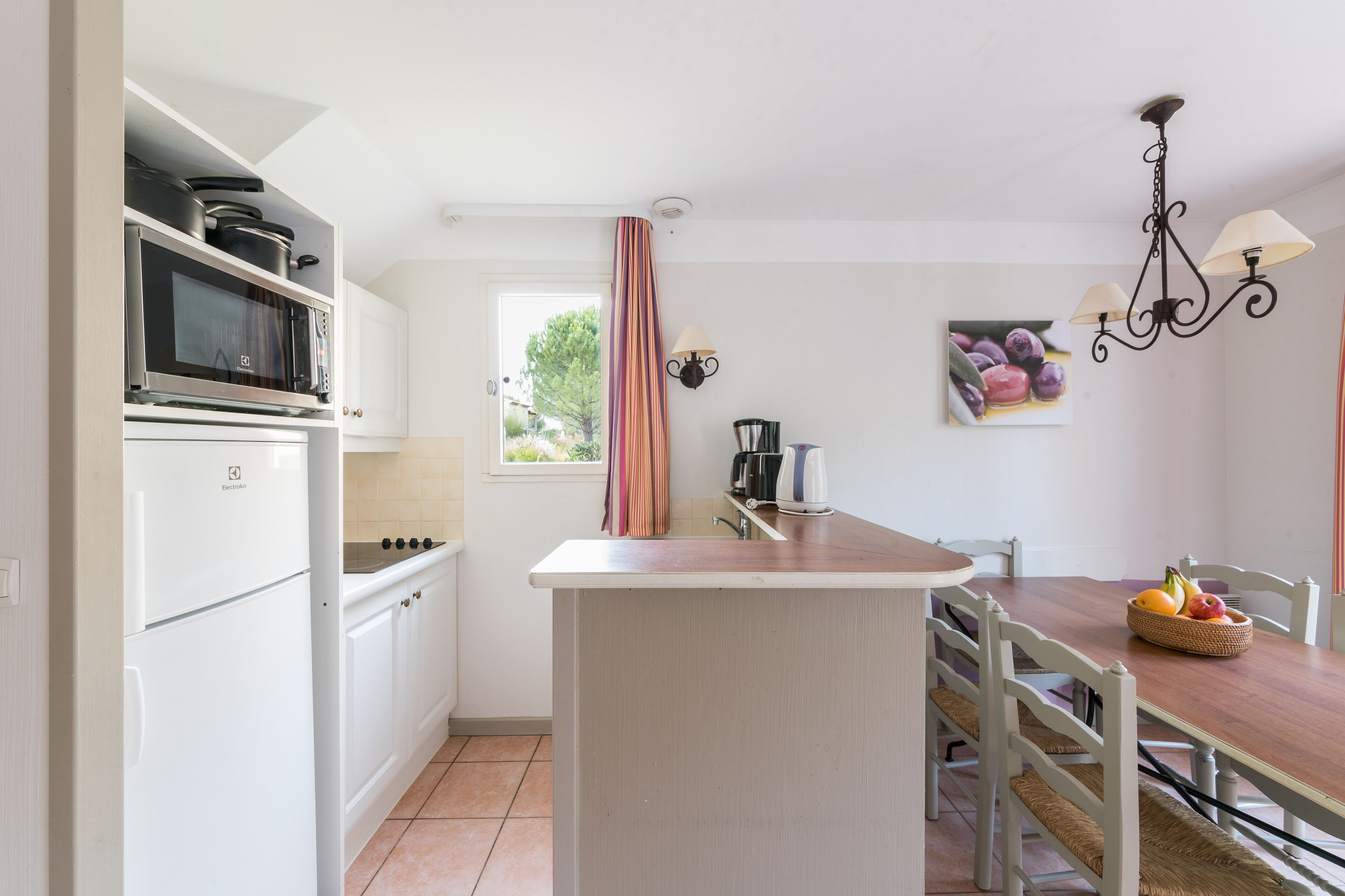 Una cocina compacta con armarios blancos, un microondas y una cafetera. Hay una mesa de comedor con sillas y una cesta de fruta.