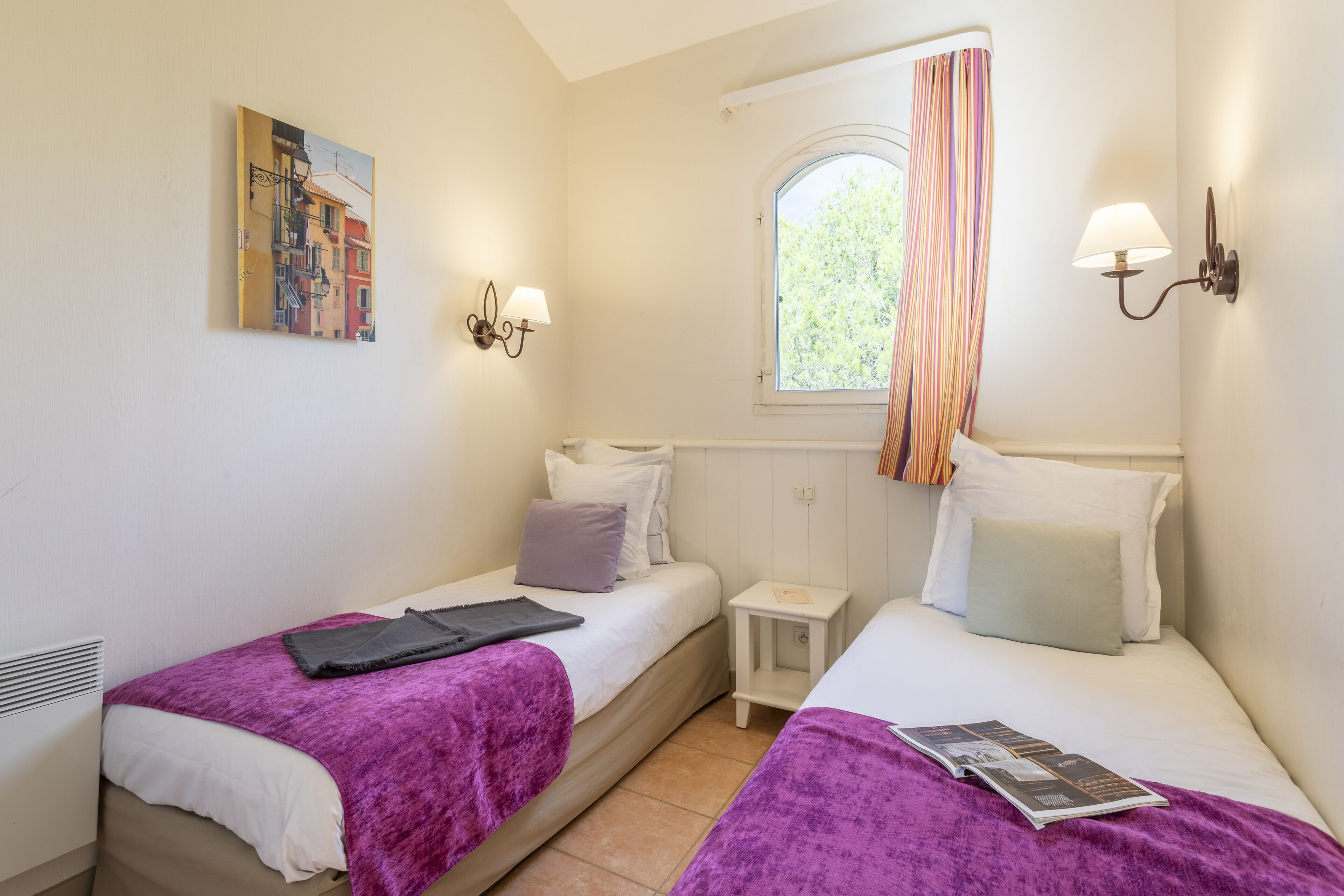 Un dormitorio con dos camas, una ventana con cortinas, una mesita de noche pequeña y lámparas de pared.