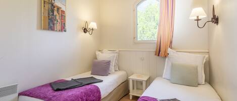 A bedroom with two beds, a window with curtains, a small bedside table, and wall-mounted lamps.