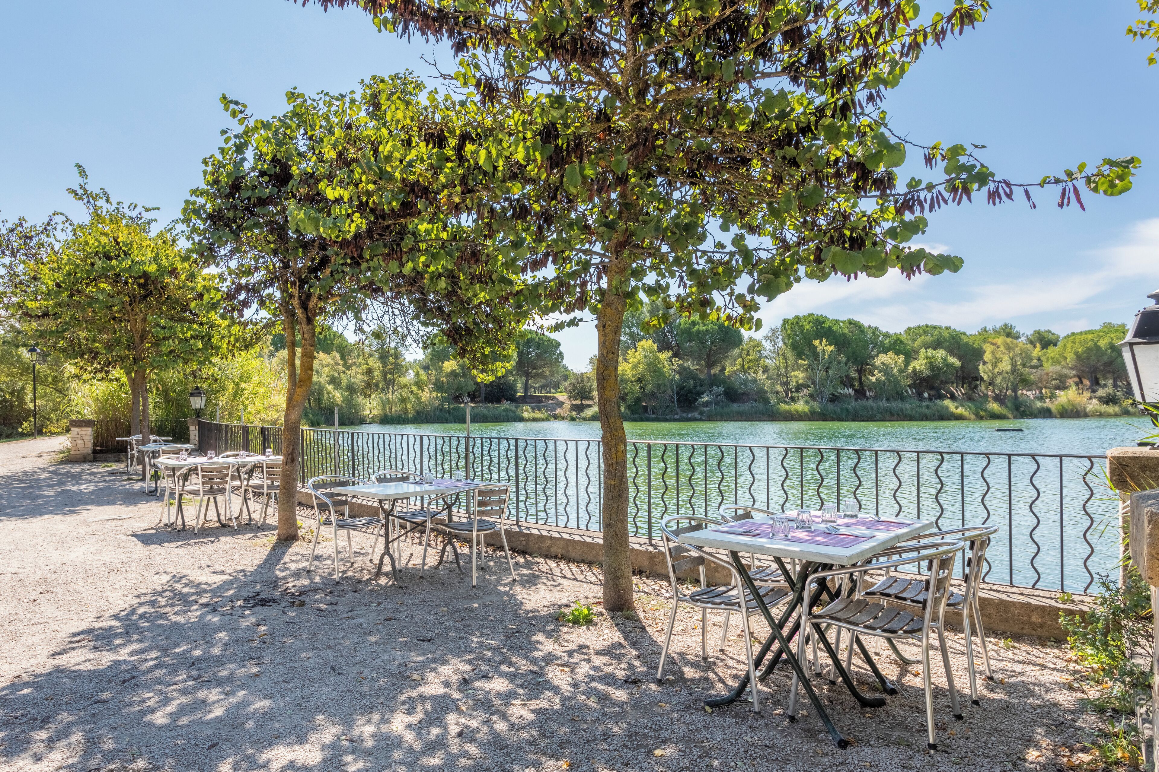 An outdoor seating area with tables and chairs, a gravel ground, and a view of a lake with trees and a fence in the background.
