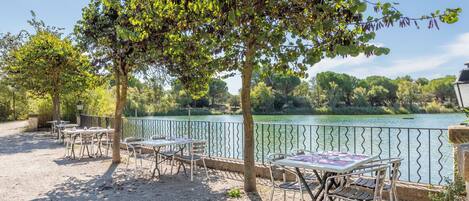 An outdoor seating area with tables and chairs, a gravel ground, and a view of a lake with trees and a fence in the background.