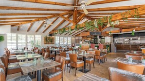 A spacious restaurant with wooden beams, large windows, and hanging plants. Tables and chairs are neatly arranged.