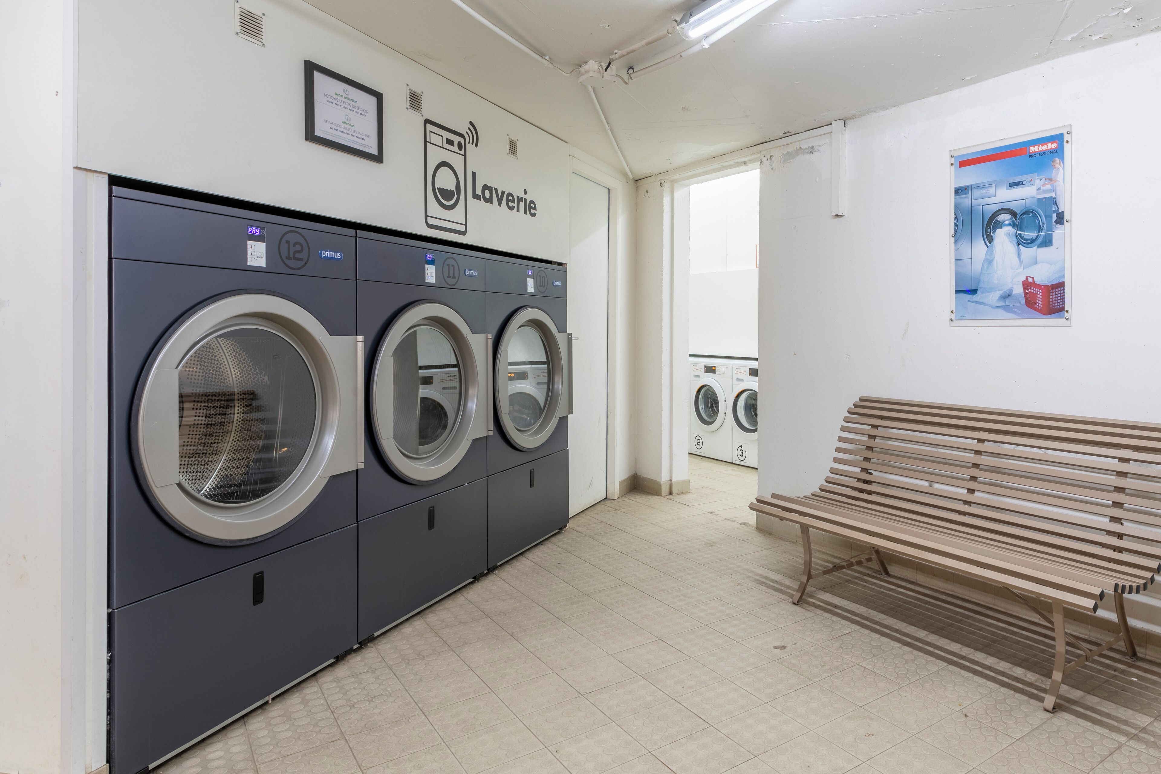 A laundry room with three front-loading washing machines, a bench, and a poster on the wall.