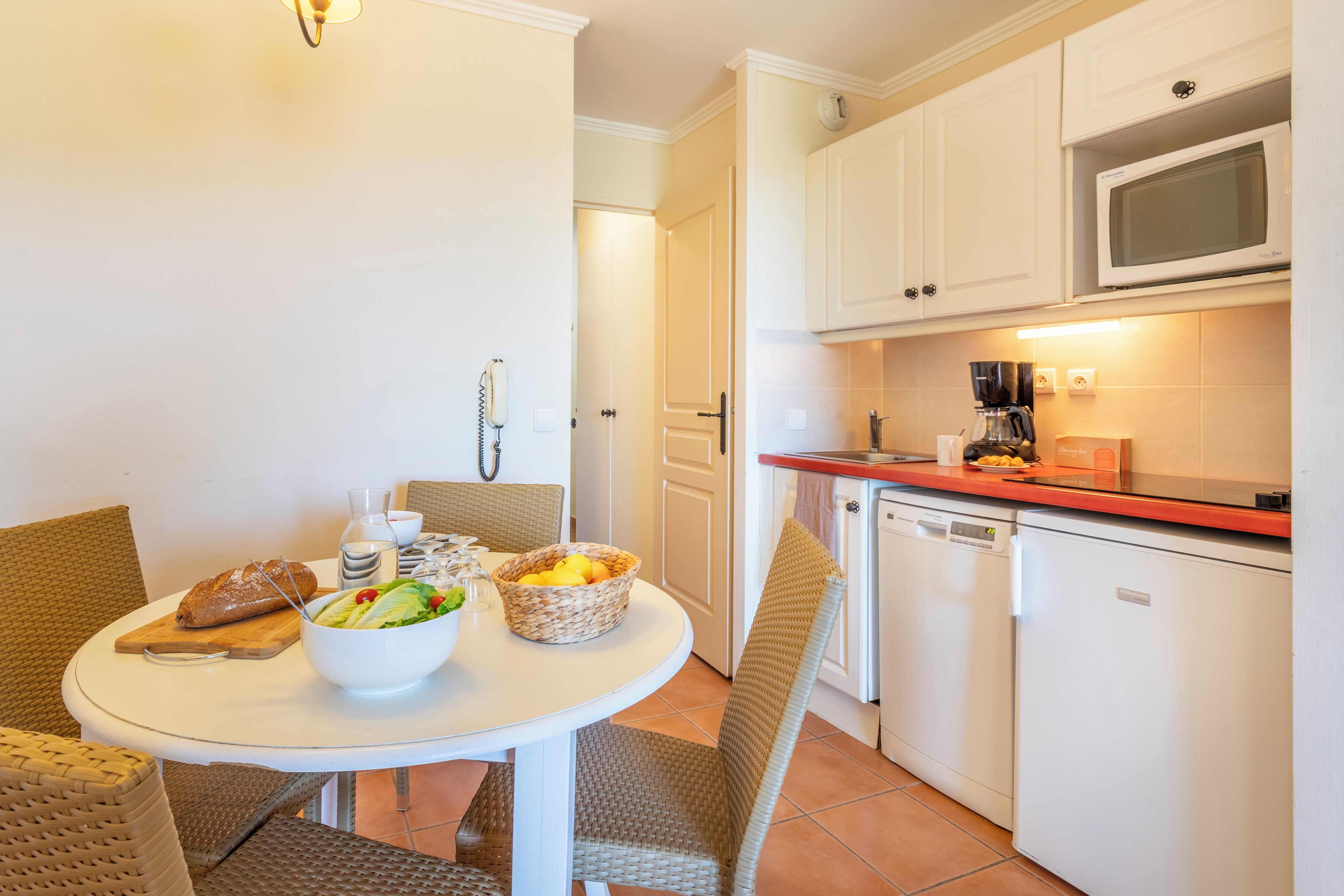 A compact kitchen with white cabinets, a microwave, and a coffee maker. There's a dining table with chairs and a bowl of salad.
