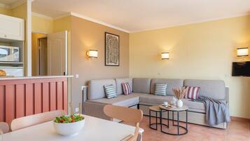 Uma sala de estar com sofá, uma mesa de jantar com cadeiras, área de cozinha com micro-ondas e uma TV fixada na parede.