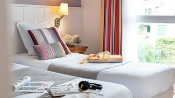 Une chambre d’hôtel avec deux lits, une lampe, un ours en peluche, une paire de lunettes et une fenêtre avec des rideaux.