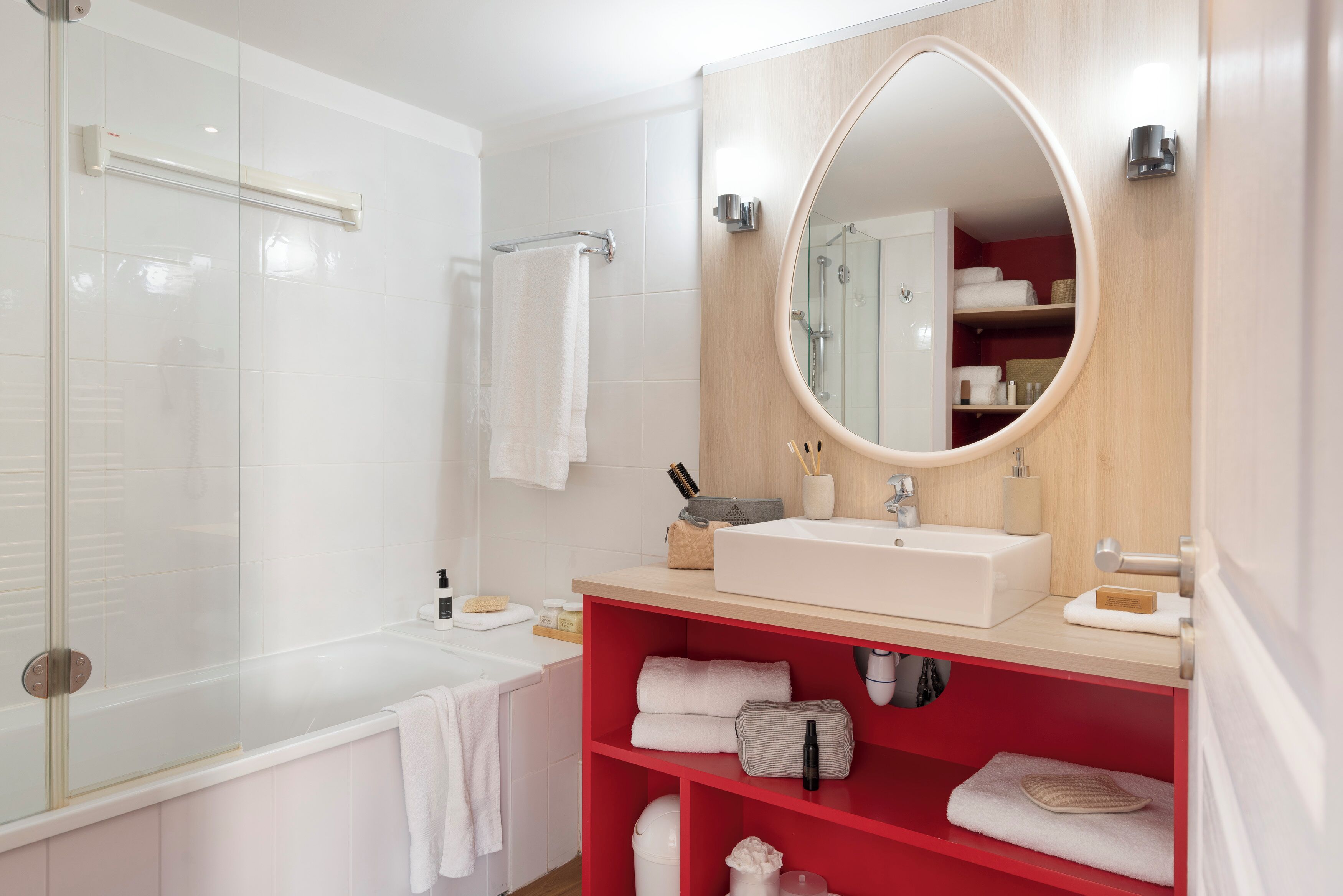 A modern bathroom with a glass-enclosed shower, a white sink with a mirror above it, and a red shelf with towels and toiletries.