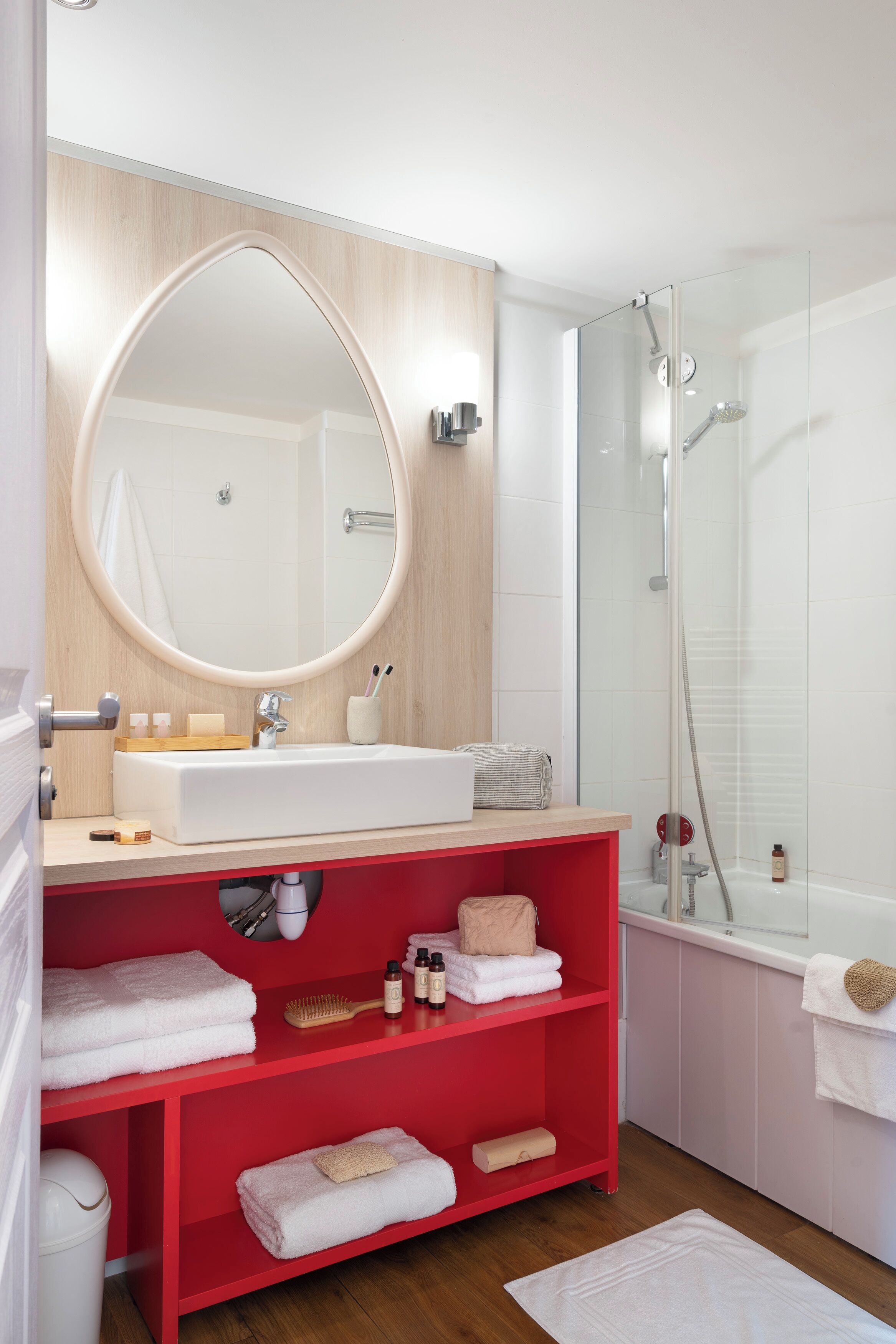Une salle de bain moderne avec un miroir rond, un lavabo blanc, des étagères rouges et une douche à parois de verre.