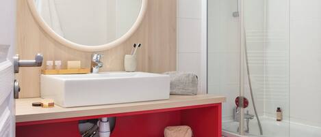 Un baño moderno con un espejo redondo, lavamanos blanco, estanterías rojas y una ducha con cerramiento de vidrio.