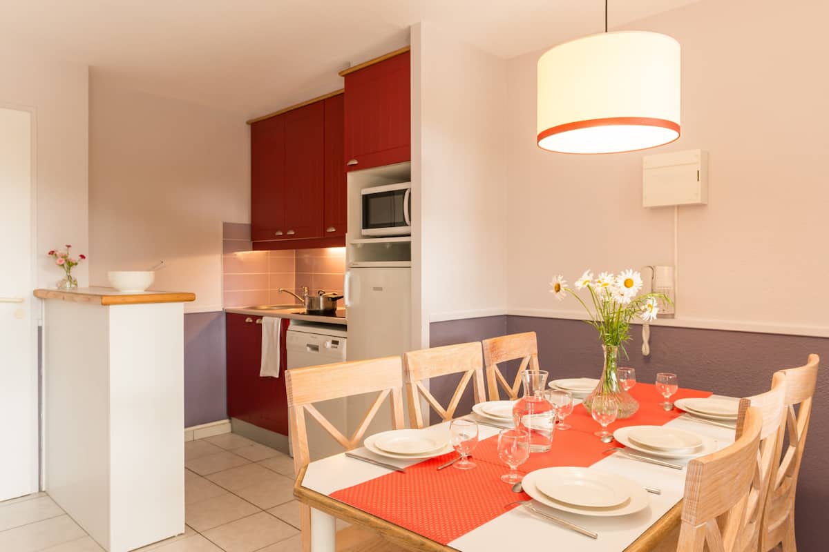 Un comedor con una mesa dispuesta para cuatro, un florero con flores y una cocina al fondo.