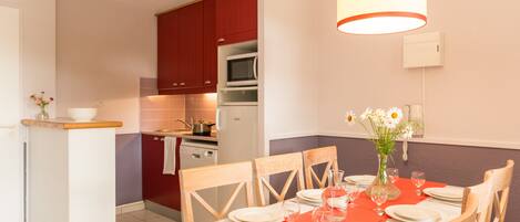 Un coin repas avec une table dressée pour quatre personnes, un vase rempli de fleurs et, au fond, une cuisine.