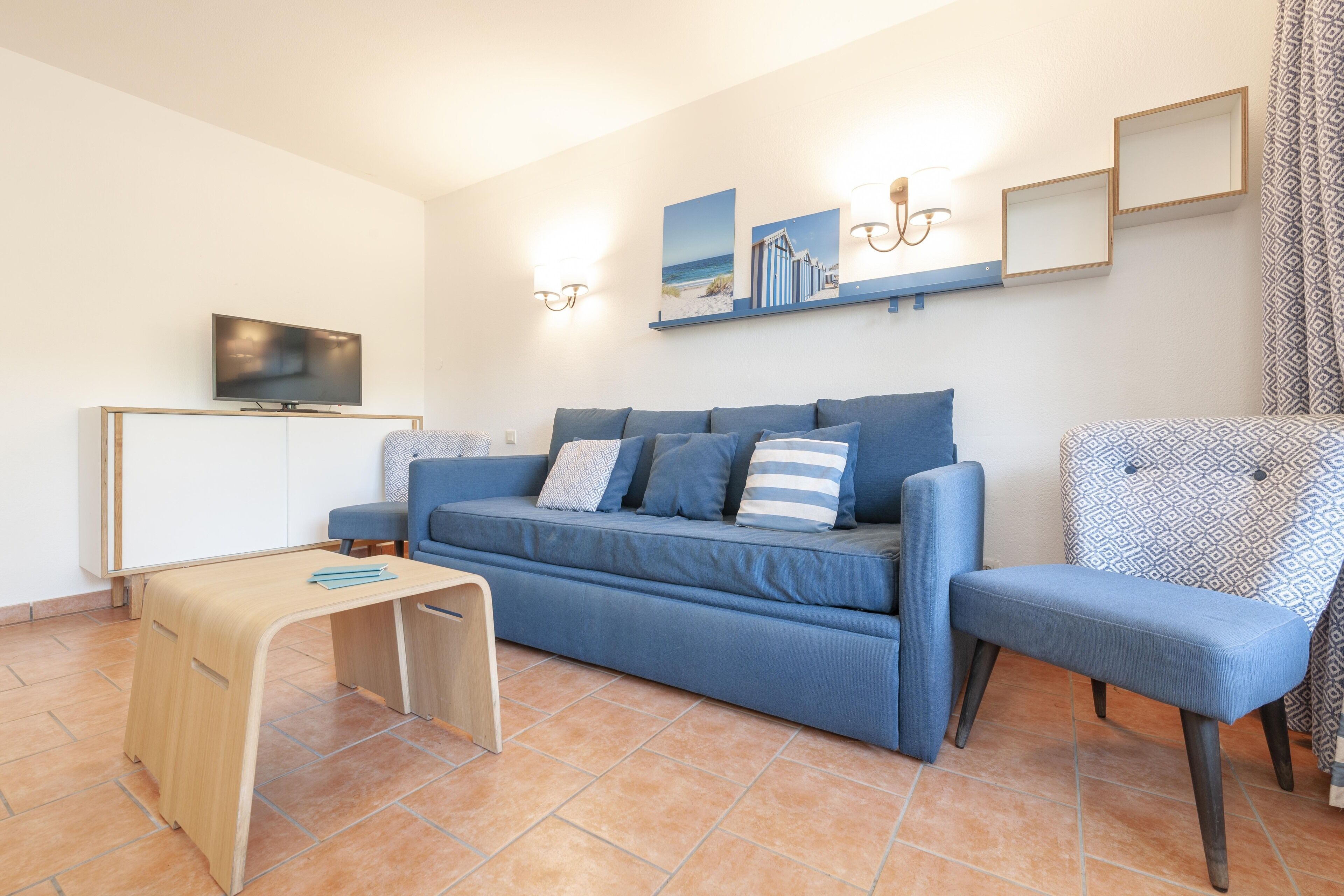 A living room with a blue sofa, a wooden coffee table, a TV on a cabinet, and wall-mounted lights.