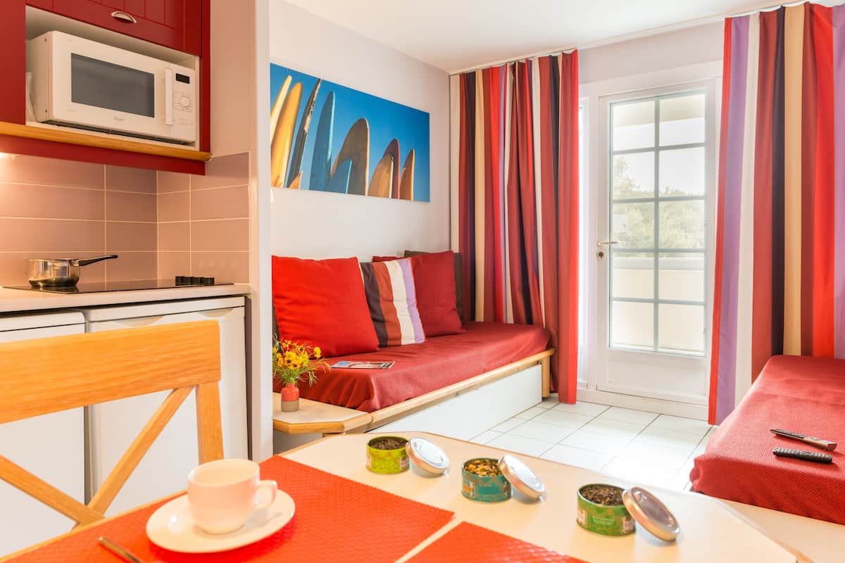 Una habitación compacta con kitchenette, una cama con ropa de cama roja, una zona de comedor con mesa y sillas, y una ventana con cortinas.