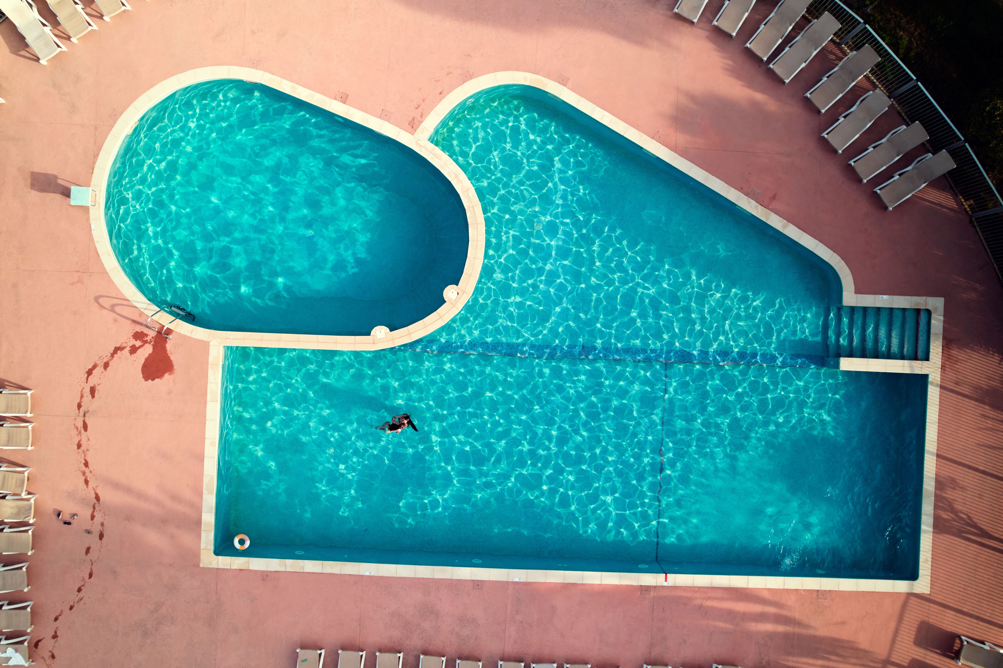 An aerial view of a swimming pool with two distinct sections, surrounded by lounge chairs and a tiled deck.