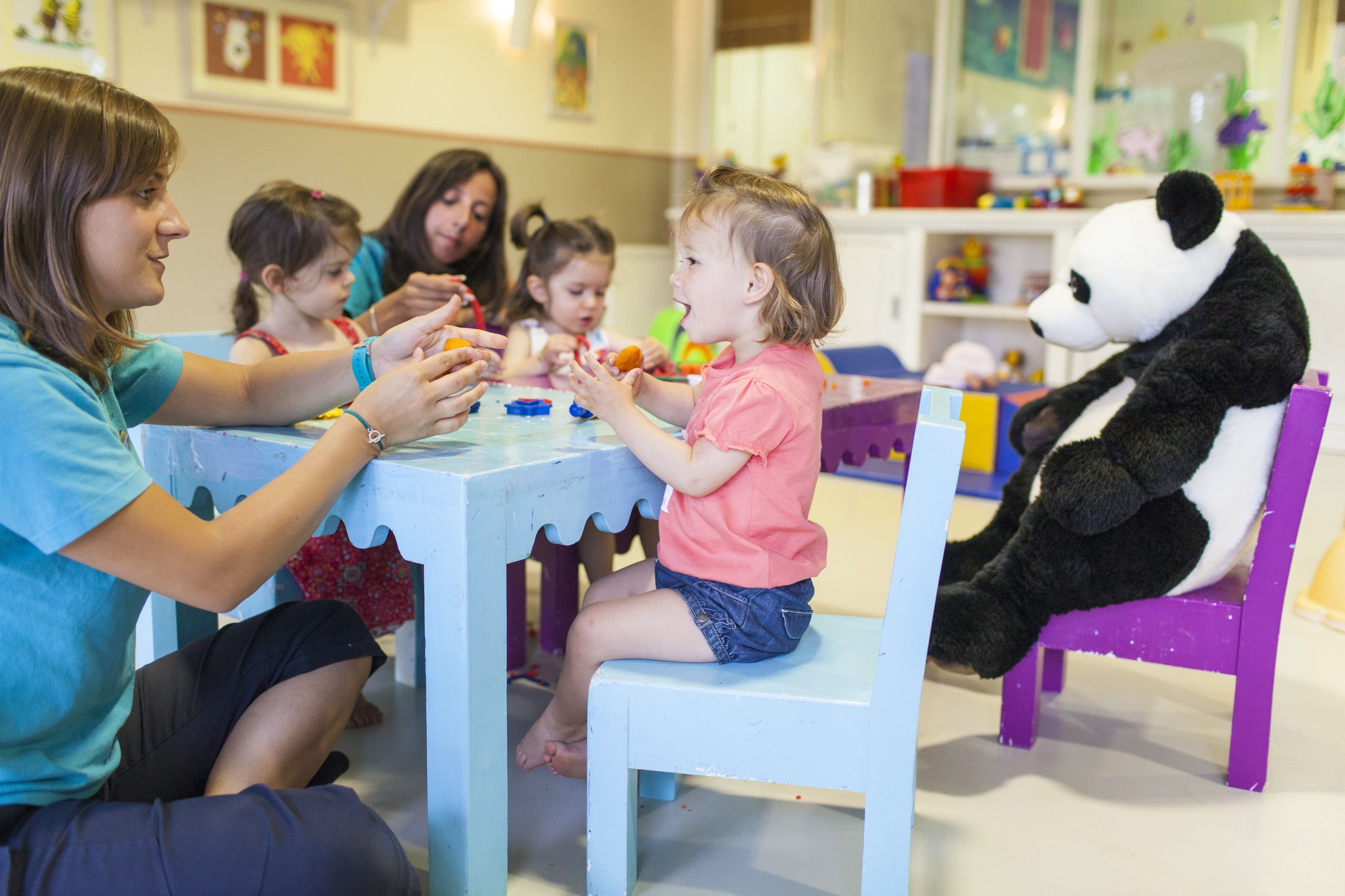 Um grupo de adultos e crianças sentados à mesa com brinquedos, em um quarto com móveis e decorações coloridas.